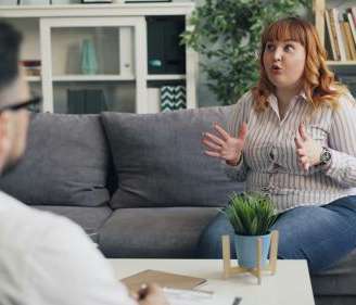 Woman sits on a couch in her therapist's office, gesturing with her hands mid-conversation. Her therapist is blurred on the left side of the photo taking notes.