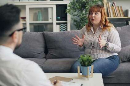 Woman sits on a couch in her therapist's office, gesturing with her hands mid-conversation. Her therapist is blurred on the left side of the photo taking notes.