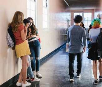 A group of teens stands inside a school hallway against the left wall, talking and looking at their phones. Another two students walk past them carrying backpacks.